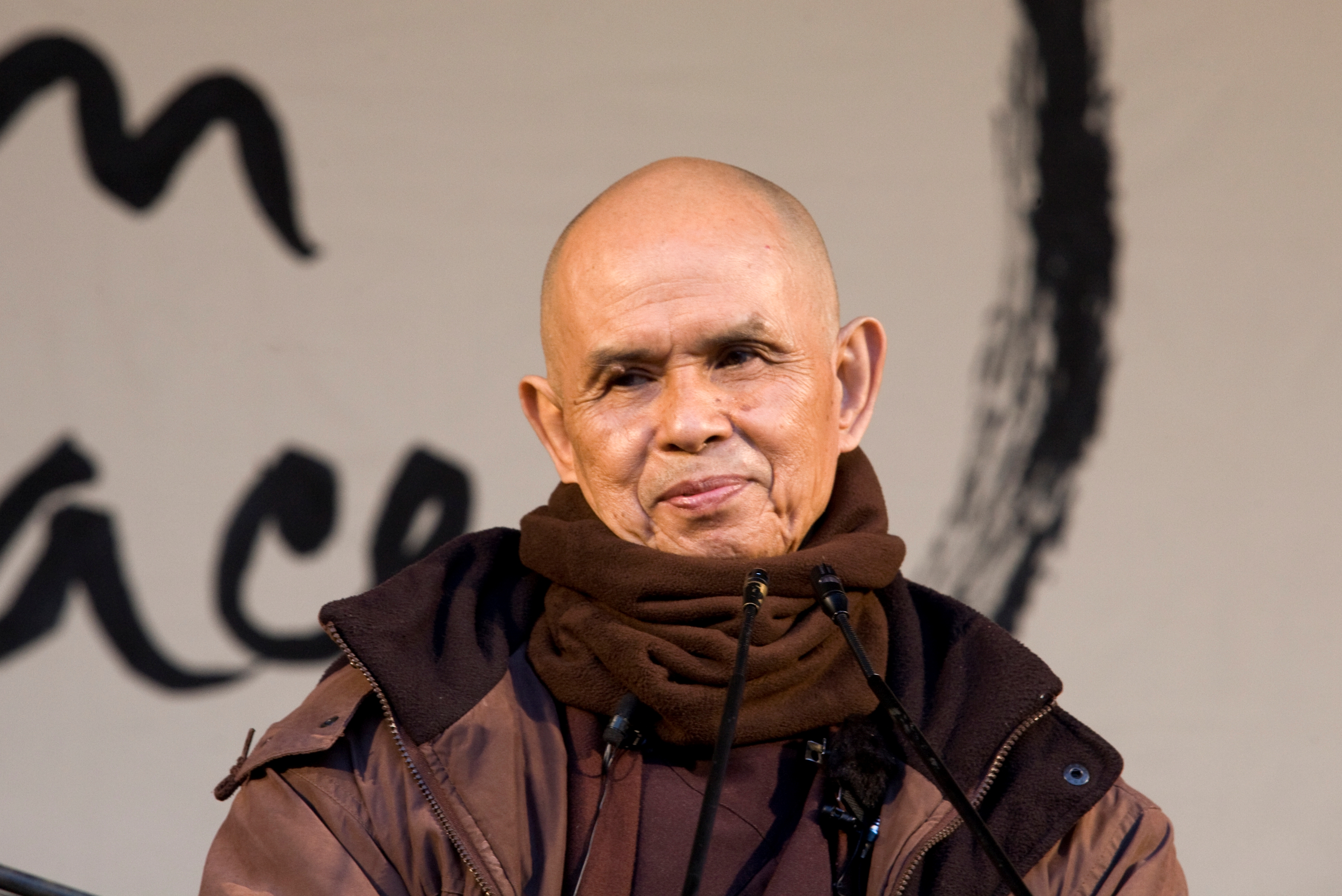 Thich Nhat Hanh warm ingepakt op een podium voor een enorme kalligrafie.